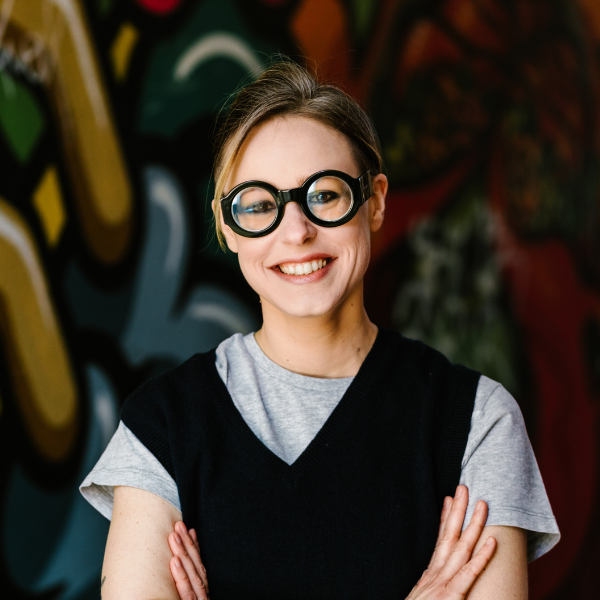 Headshot of a white middle-aged woman with brown hair tied at the back and statement round glasses wearing a grey T-shirt and a black waistcoat